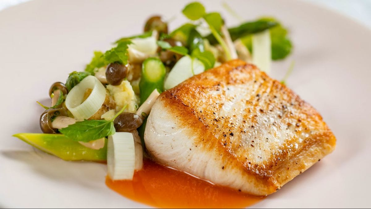 A seared white fish fillet served on a light plate with sautéed mushrooms, sliced leeks, asparagus, and a vibrant orange sauce drizzled underneath at one of the Disney World Michelin Restaurants