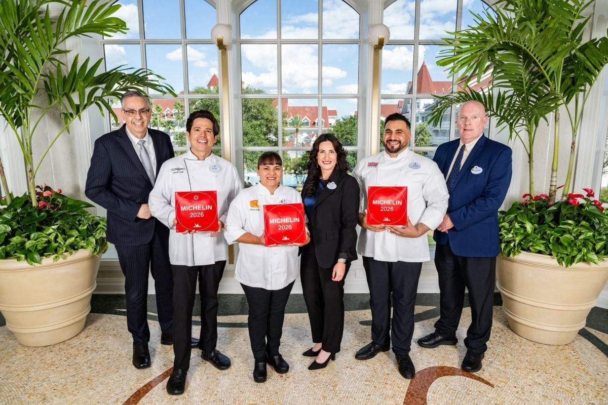 Five people stand in a bright, sunlit room, with two chefs holding red Walt Disney World Michelin Restaurant plaques, flanked by three professionals in formal business attire.